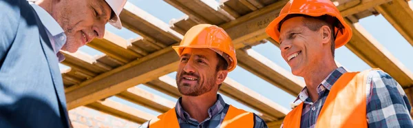 Panoramaaufnahme von Bauarbeitern in der Nähe eines gestandenen Geschäftsmannes mit Helm — Stockfoto