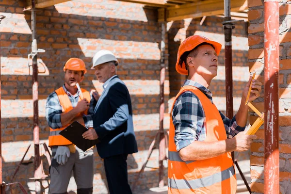 Selective focus of mature constructor measuring building near coworker and businessman in helmets — Stock Photo
