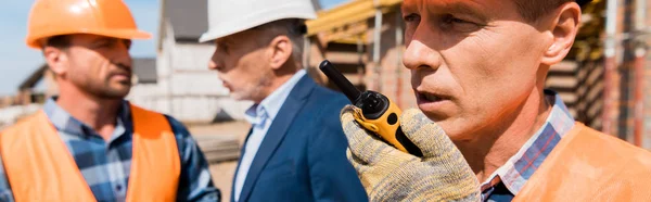 Colpo panoramico del costruttore che tiene walkie talkie mentre parla vicino a collega e uomo d'affari — Foto stock