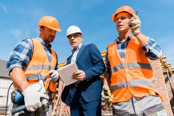 Низкий угол зрения строителя держит рацию Talkie рядом с коллегой и бизнесменом с цифровым планшетом — стоковое фото