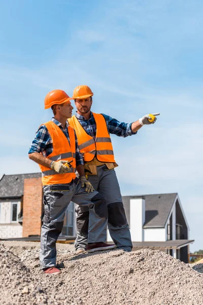 Hermoso constructor apuntando con el dedo mientras está de pie sobre piedras cerca de compañero de trabajo - foto de stock