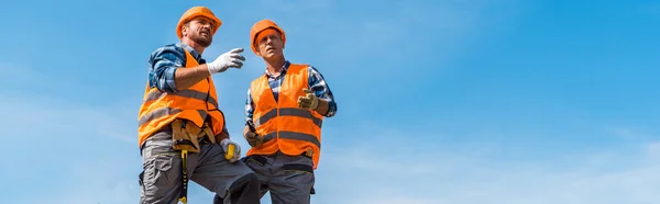 Tiro panorâmico de colegas de trabalho em capacetes laranja em pé sobre rochas contra o céu — Stock Photo