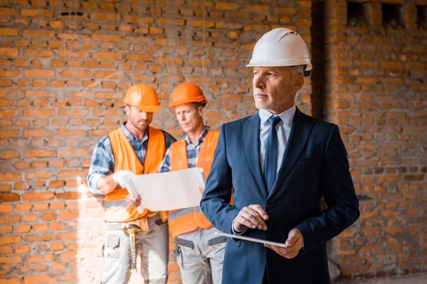 Selective focus of mature businessman holding digital tablet near constructors — Stock Photo