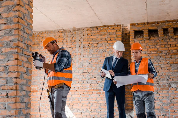Handsome builder using hammer drill near businessman and coworker — Stock Photo