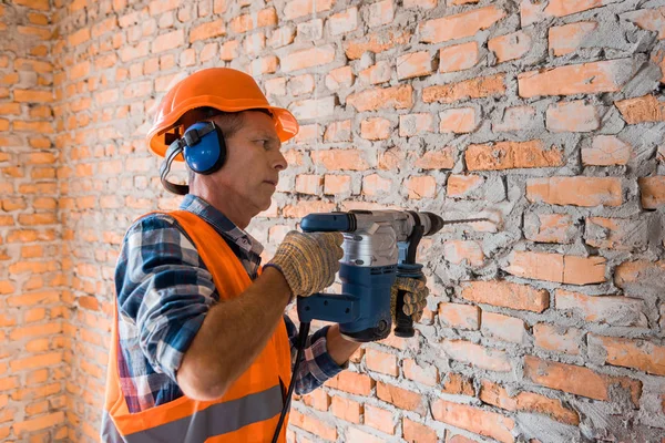 Construtor maduro no capacete usando martelo broca perto da parede de tijolo — Fotografia de Stock