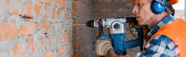 Panoramic shot of mature builder using hammer drill near brick wall — Stock Photo