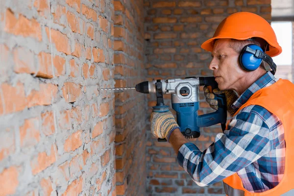 Constructor maduro utilizando taladro de martillo cerca de la pared de ladrillo - foto de stock