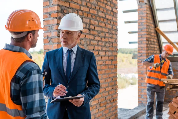 Hombre de negocios barbudo en casco sujetando portapapeles cerca de constructores - foto de stock