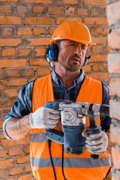 Enfoque selectivo de constructor guapo y barbudo que sostiene taladro de martillo cerca de la pared de ladrillo - foto de stock