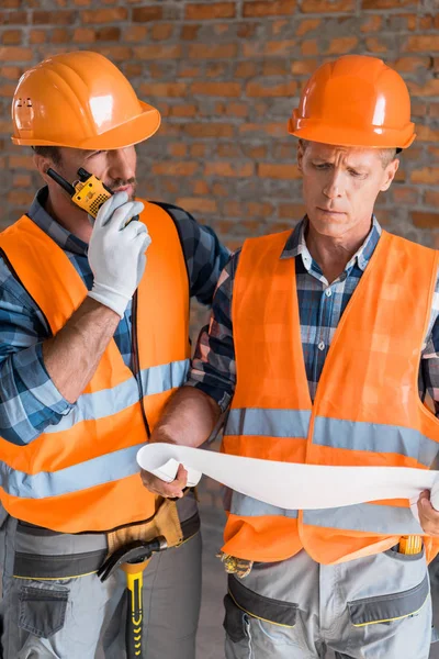 Mature constructor holding blueprint near coworker with walkie talkie — Stock Photo