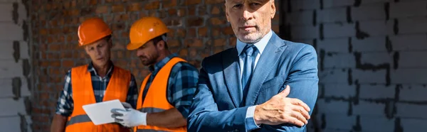 Tiro panorâmico de empresário de pé com braços cruzados perto de construtores — Stock Photo