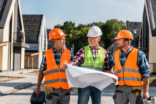 Constructor maduro sosteniendo plano cerca de compañeros de trabajo y casas - foto de stock