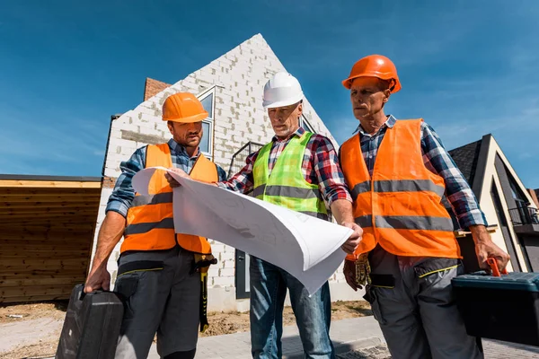 Bauarbeiter in Helmen halten Werkzeugkästen in der Nähe von Häusern und betrachten Baupläne — Stockfoto
