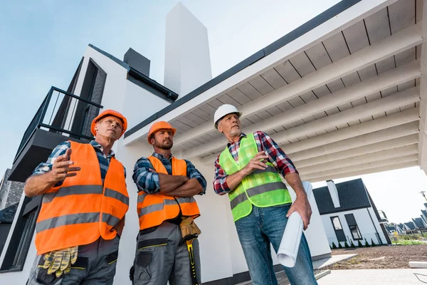 Low angle view of constructors in helmets standing near coworker with blueprint — Stock Photo