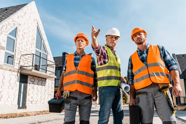 Construtores em capacetes segurando caixas de ferramentas perto de colegas de trabalho gestos perto de casas — Fotografia de Stock
