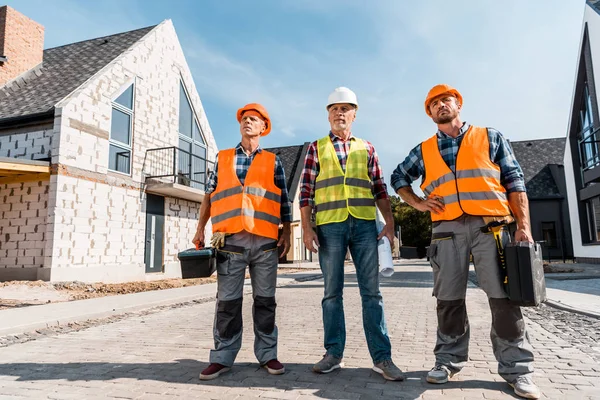 Constructores de mediana edad en cascos de pie con compañero de trabajo cerca de casas - foto de stock