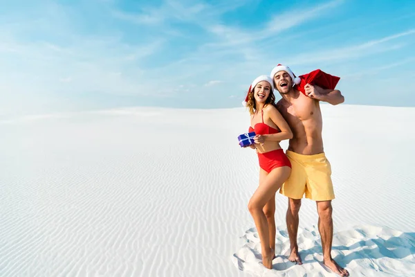 Сексуальная и улыбающаяся девушка с подарком и парнем с мешком Санты на пляже на Мальдивах — стоковое фото