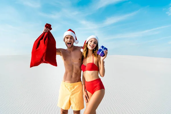 Sexy e sorridente namorada segurando presente e namorado com santa saco na praia em Maldivas — Fotografia de Stock