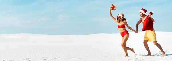 Panoramic shot of sexy and smiling girlfriend holding gift and boyfriend with santa sack on beach in Maldives — Stock Photo