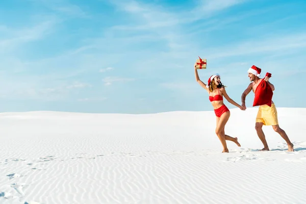 Сексуальная и улыбающаяся девушка с подарком и парнем с мешком Санты на пляже на Мальдивах — стоковое фото