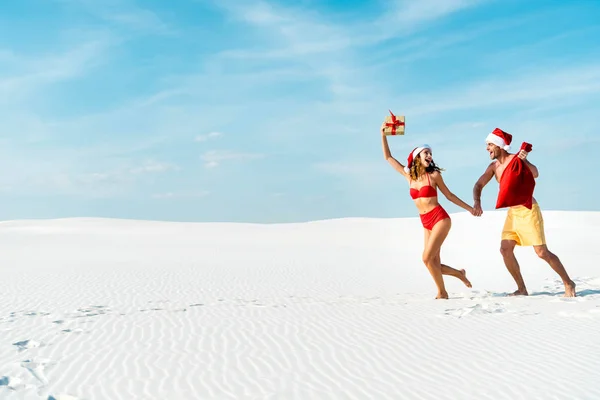 Sexy y sonriente novia sosteniendo regalo y novio con santa saco en la playa en Maldivas - foto de stock