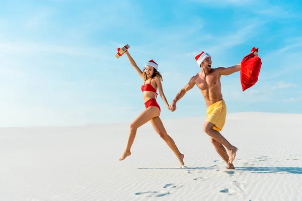 Sexy y sonriente novia sosteniendo regalo y novio con santa saco corriendo en la playa en Maldivas - foto de stock