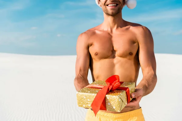 Обрезанный вид сексуального мужчины в шляпе Санты с подарком на пляже на Мальдивах — стоковое фото