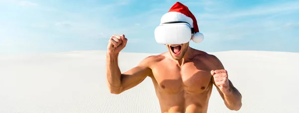 Tiro panorámico de hombre sexy en sombrero de santa con auriculares de realidad virtual que muestra sí gesto en la playa en Maldivas — Stock Photo