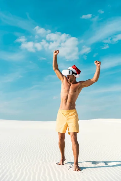 Sexy hombre en sombrero de santa con auriculares de realidad virtual que muestra sí gesto en la playa en Maldivas - foto de stock
