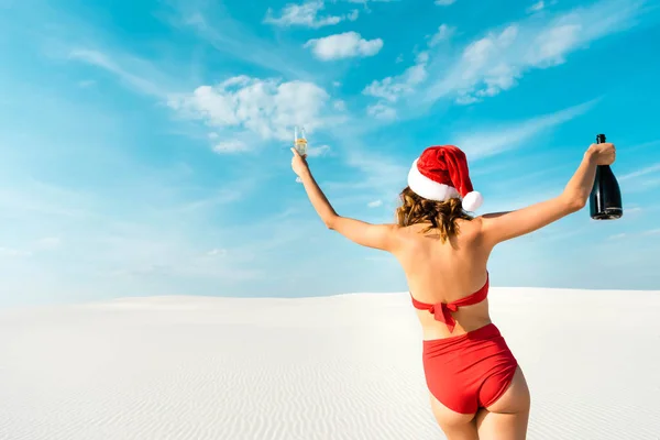 Vista trasera de la mujer sexy en sombrero de santa y traje de baño celebración de copa de champán y botella en la playa en Maldivas - foto de stock