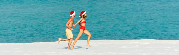 Panoramic shot of sexy girlfriend and boyfriend in santa hats running on beach in Maldives — Stock Photo