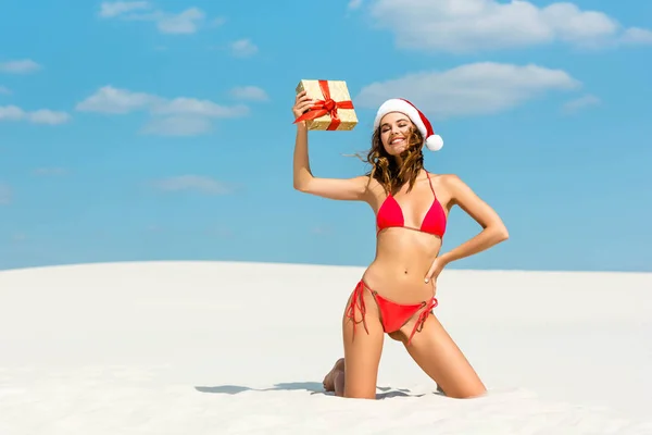 Sexy and smiling woman in santa hat and swimsuit holding christmas gift on beach in Maldives — Stock Photo