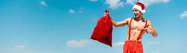 Panoramic shot of handsome and sexy man in santa hat pointing with finger at santa sack on beach in Maldives — Stock Photo