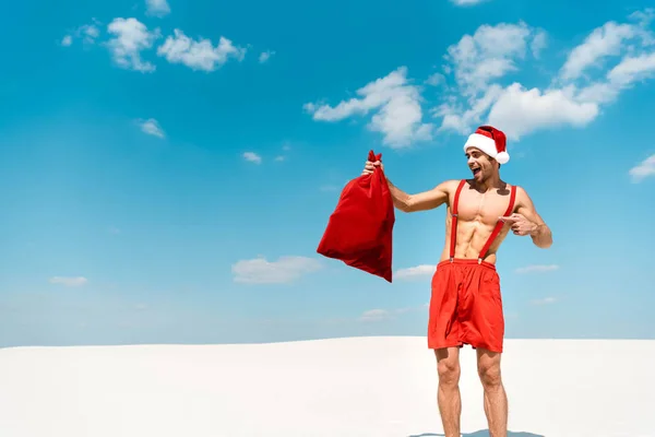 Uomo bello e sexy in cappello di Babbo Natale che punta con il dito al sacco di Babbo Natale sulla spiaggia delle Maldive — Foto stock