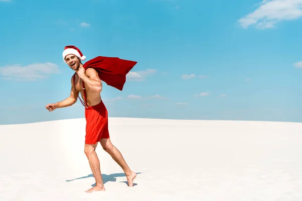 Handsome and sexy man in santa hat holding santa sack and walking on beach in Maldives — Stock Photo