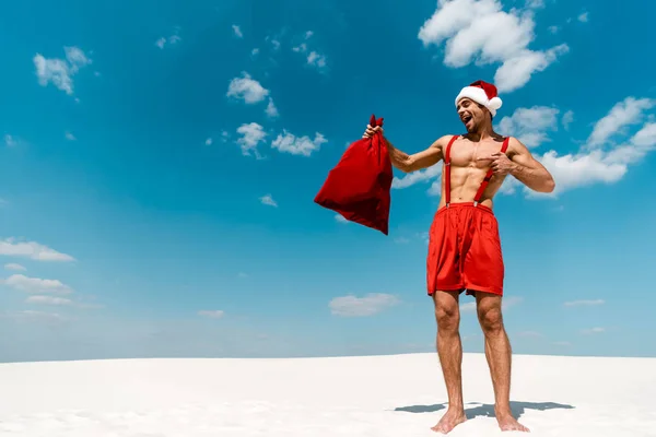 Uomo bello e sexy in cappello di Babbo Natale che punta con il dito al sacco di Babbo Natale sulla spiaggia delle Maldive — Foto stock