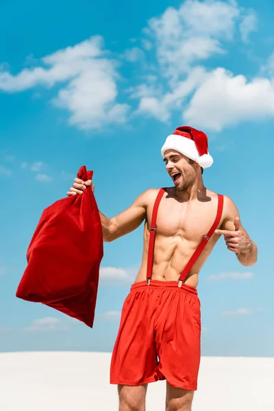 Uomo bello e sexy in cappello di Babbo Natale che punta con il dito al sacco di Babbo Natale sulla spiaggia delle Maldive — Foto stock