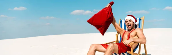 Plano panorámico de hombre guapo y sexy en sombrero de santa celebración saco de santa y sentado en la silla de cubierta en la playa en Maldivas - foto de stock