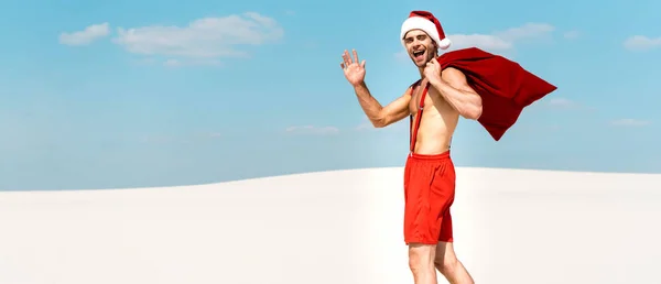 Panoramic shot of handsome and sexy man in santa hat holding santa sack and waving on beach in Maldives — Stock Photo