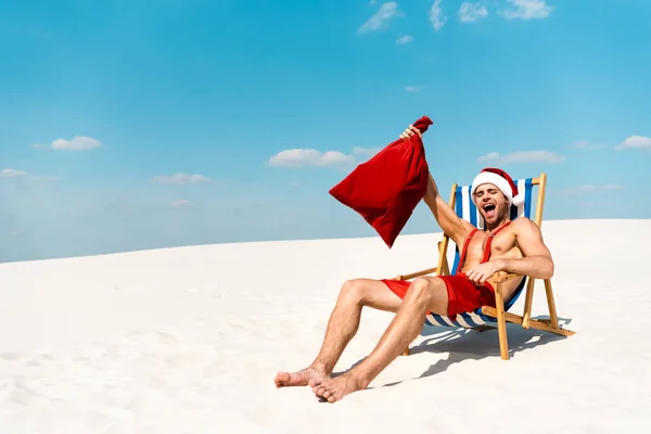 Handsome and sexy man in santa hat holding santa sack and sitting on deck chair on beach in Maldives — Stock Photo