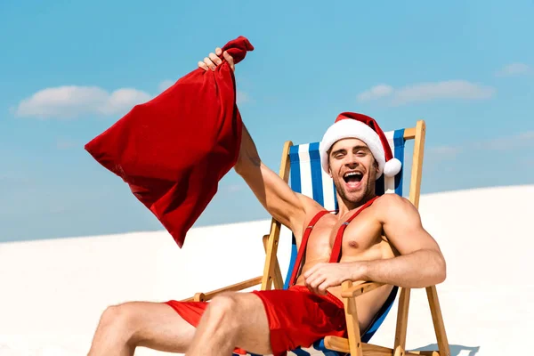 Handsome and sexy man in santa hat holding santa sack and sitting on deck chair on beach in Maldives — Stock Photo