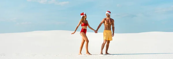 Panoramic shot of sexy girlfriend and boyfriend in santa hats holding hands and walking on beach in Maldives — Stock Photo