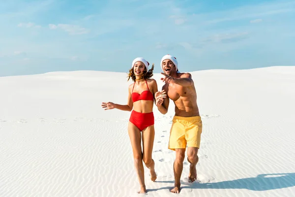 Sexy novia y novio en santa sombreros corriendo y señalando el dedo en la playa en Maldivas - foto de stock