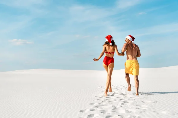 Back view de namorada sexy e namorado em chapéus santa correndo na praia em Maldivas — Fotografia de Stock