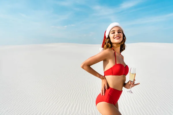 Sexy e sorridente donna in cappello di Babbo Natale e costume da bagno in possesso di bicchiere di champagne sulla spiaggia in Maldive — Foto stock