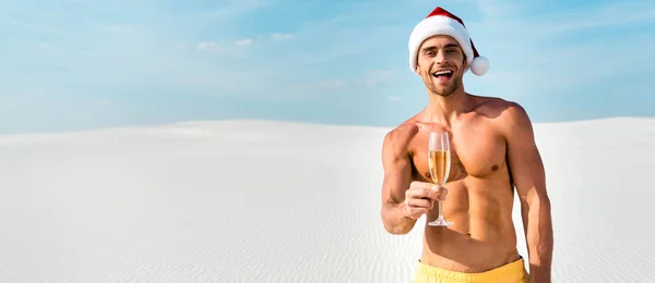 Panoramic shot of sexy man in santa hat holding champagne glass on beach in Maldives — Stock Photo
