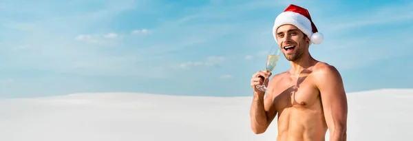 Panoramic shot of sexy man in santa hat holding champagne glass on beach in Maldives — Stock Photo