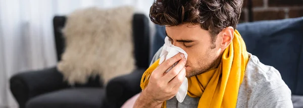 Panoramic shot of sick man in scarf sneezing and holding napkin — Stock Photo