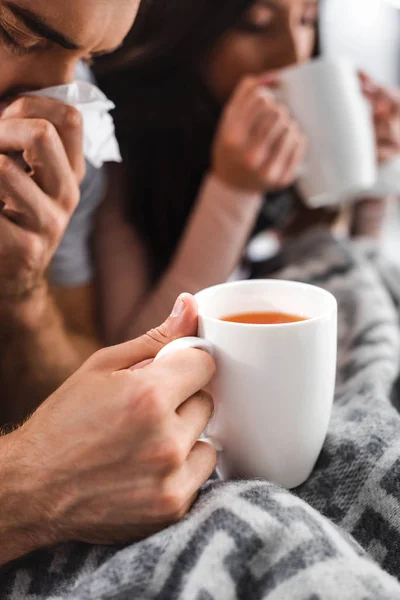 Cropped view of sick girlfriend and boyfriend holding cups and sneezing with tea — Stock Photo