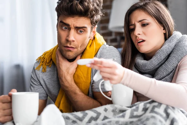 Enfoque selectivo de novia enferma y novio sosteniendo tazas y mirando el termómetro - foto de stock